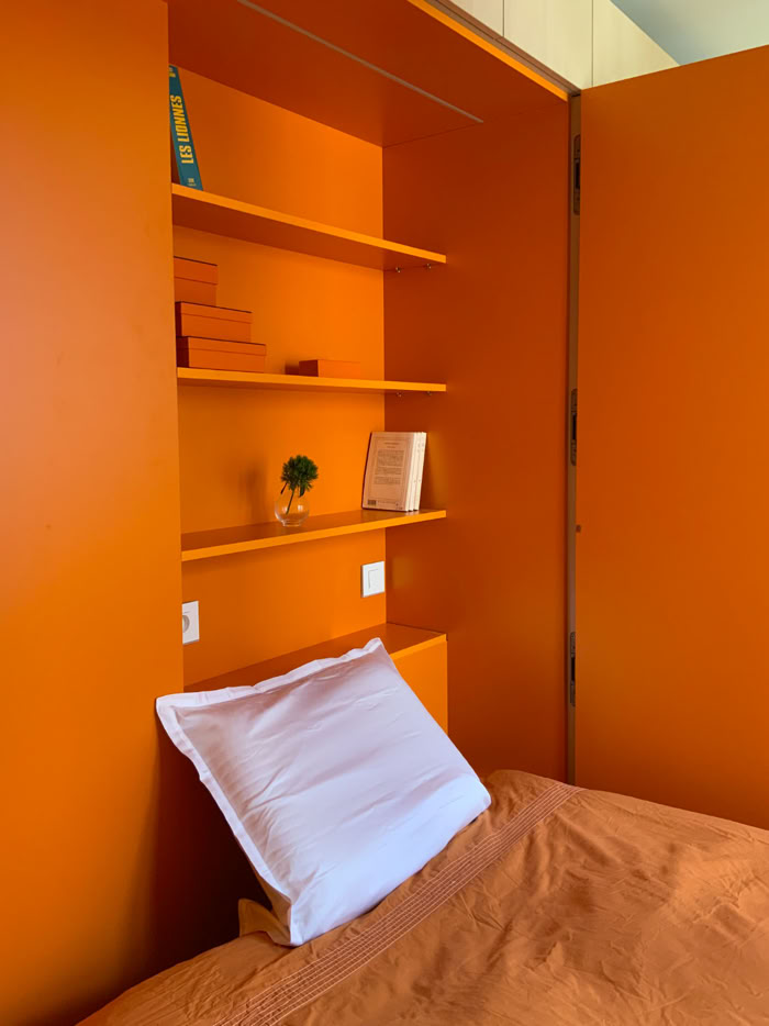 Zoom sur l'espace nuit orange vif avec tête de lit et étagères intégrées dans un studio de 17 m².